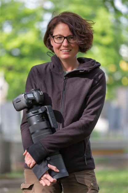Porträt der Fotografin Perdita Petzl. Die Forografin hat eine Kamera mit einem großen Objektiv im Arm. Im Hintergrund ein Park und Bäume.