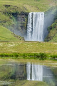 Auf dem Bild ist eine Grüne Berglandschaft zu sehen mit einem Wasserfall. Im Vordergrund fließt ein Fluss entlang. Islands Süden im Spätsommer.