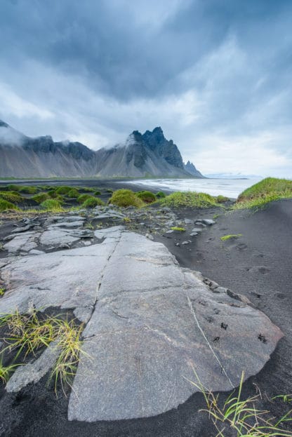 Schöne Landschaftsaufnahme auf Island. Im Vordergrund ist der schwarze Strand mit Steinen und mit grünem Gras bewachsen zu sehen. Im Hintergrund die Berge, die teilweise von Wolken verdeckt werden. Islands Süden im Spätsommer. Vulkan, Landschaft und Eis!