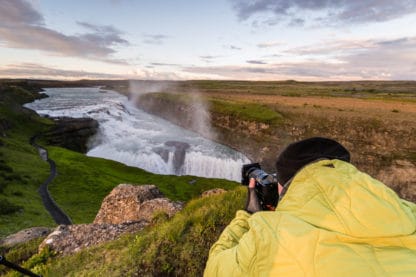 Ein Fotograf mit einer gelben Jacke hält seine Kamera in der Hand und fotografiert einen Gletscher. Links davon eine grüne Wiese mit einem schwarzen Pfad. Rechts davon braune Felsen. Islands Süden im Spätsommer. Landschaftsbild.