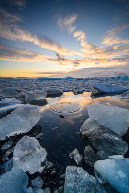 Eisige Landschaft in Island. Im Vordergrund große Eisstücke auf dem Wasser das Kreise zieht. Im Hintergrund Berge und der Himmel in Abendstimmung. Blauer Himmel und gelblich angestrahlte Wolken. Islands Süden im Spätsommer. Landschaft und Eis.