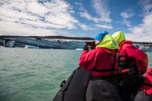 Zwei Fotografen in roten Jacken machen eine Expedition auf einem großen Schlauchboot. Sie fotografieren die Landschaft. Im Vordergrund sieht man Wasser, im Hintergrund Gletscher, Schnee und den blauen aber bewölkten Himmel. Islands Süden im Spätsommer. Eisige Landschaft.