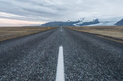 Der Betrachter des Bildes sitzt auf dem Mittelstreifen einer langen Straße in Island. Rechts und links von der Straße trockene Gräser. Im Hintergrund große Felsen mit Schnee bedeckt. Der Himmel ist grau. Islands Süden im Spätsommer. Landschaftsbild.