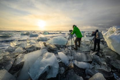 Zwei Fotografen stehen mit ihren Stativen und Kameras zwischen großen Eisstücken auf einem Gletscher und fotografieren die untergehende Sonne. Islands Süden im Spätsommer. Vulkan, Landschaft und Eis.