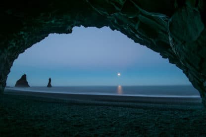 Auf dem Foto ist ein Felsbogen zu sehen, durch den der Betrachter durchschaut auf das Meer in der Nacht. Der Himmel ist blau, der Mond geht gerade auf und auf der linken Seite stehen zwei schwarze Felden im Wasser. Islands Süden im Spätsommer.