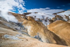 Es ist ein Landschaftsbild aus Island zu sehen. Im Vordergrund braune Berge, im Hintergrund liegt an den Hängen Schnee. Der Himmel ist blau und man sieht weiße Wolken. Islands Süden im Spätsommer. Vulkan, Landschaft und Eis.