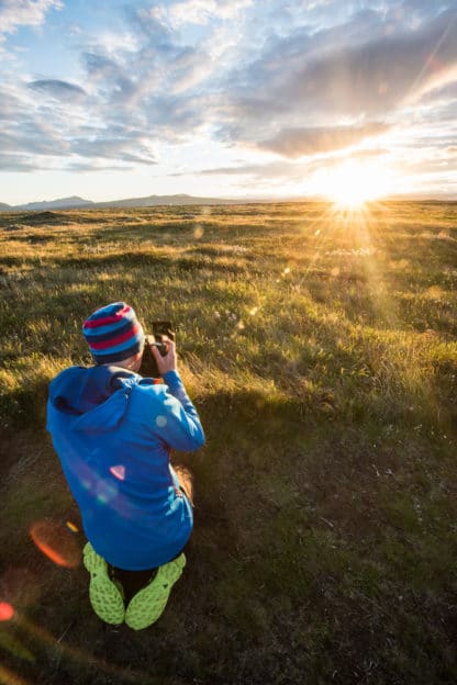 Ein Fotograf mit einer blauen Jacke und seiner Kamera sitzt in Island auf einer Wiese. Die Sonne geht gerade unter, das Gras wird angestrahlt und der Himmel ist blau und bewölkt. Im fernen Hintergrund sieht man ein paar Berge. Islands Süden im Spätsommer. Vulkan, Landschaft und Eis!