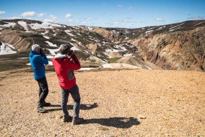Zwei Fotografen in Island fotografieren die Landschaft - Felsen die mit Schnee und Gras bedeckt sind. Der Himmel ist blau. Die zwei Personen tragen blaue und rote Jacken. Islands Süden im Spätsommer. Vulkan, Landschaft und Eis!